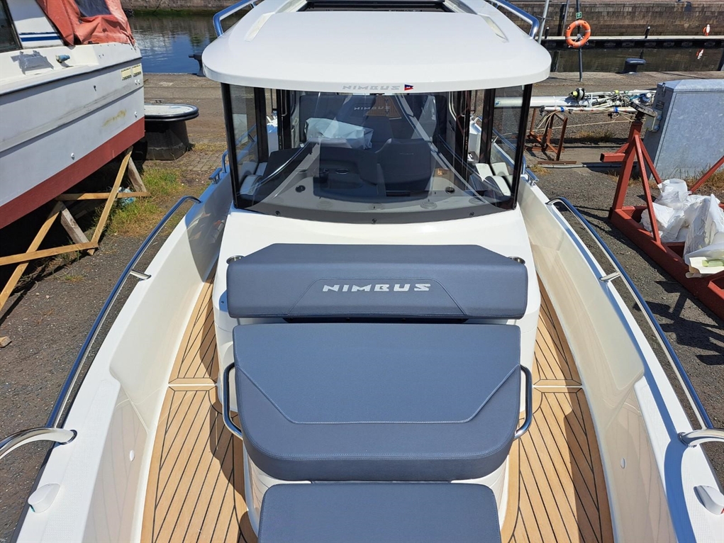 Nimbus boat interior with a sunbathing area, blue cushions, and a gleaming wooden deck by the water, showcasing nautical design.